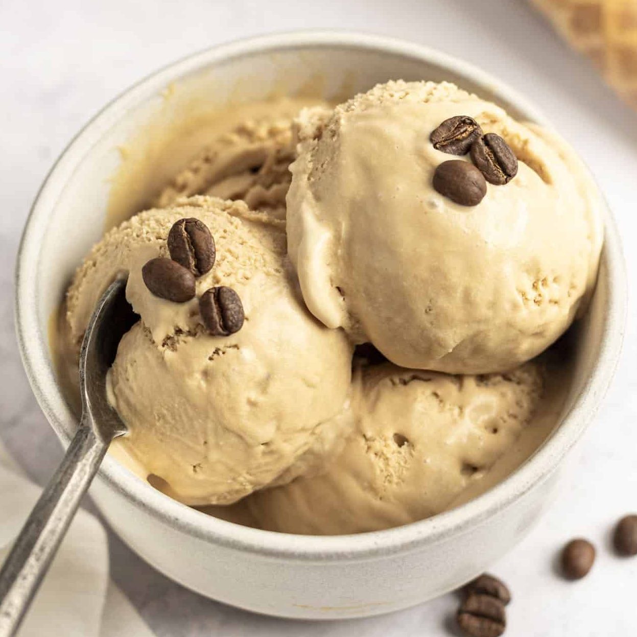 Creamy coffee-flavored ice cream scoops in a white bowl, topped with coffee beans, and a spoon resting on the edge.