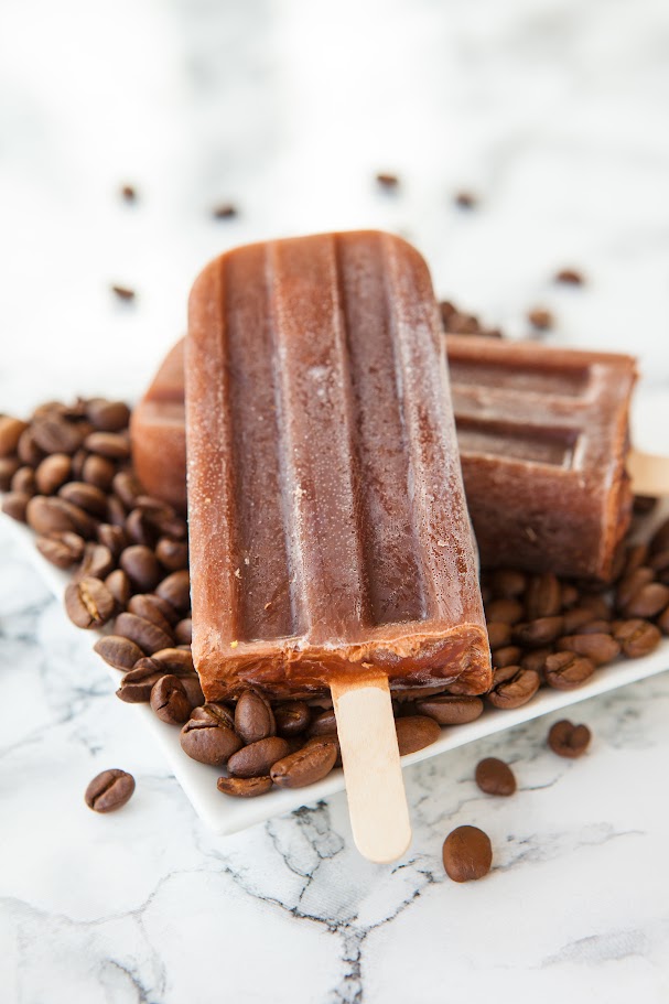 Two chocolate coffee popsicles on a plate surrounded by coffee beans, set on a marble surface. Refreshing treat for coffee lovers.